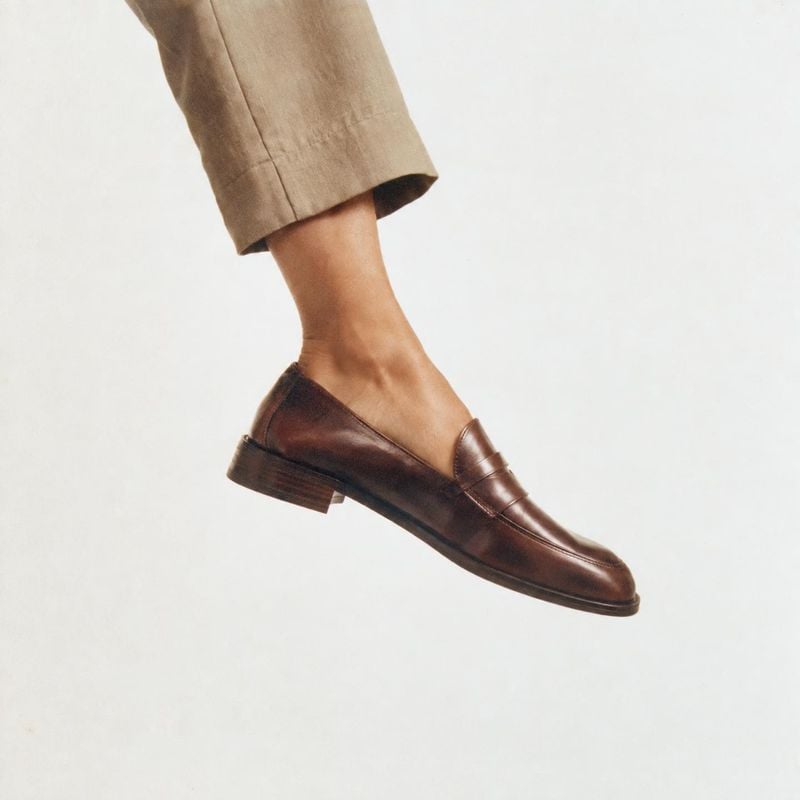 brown leather loafer on-body detail product photo: A cropped model wears a polished brown leather loafer with tailored beige trousers against a bright, minimal…