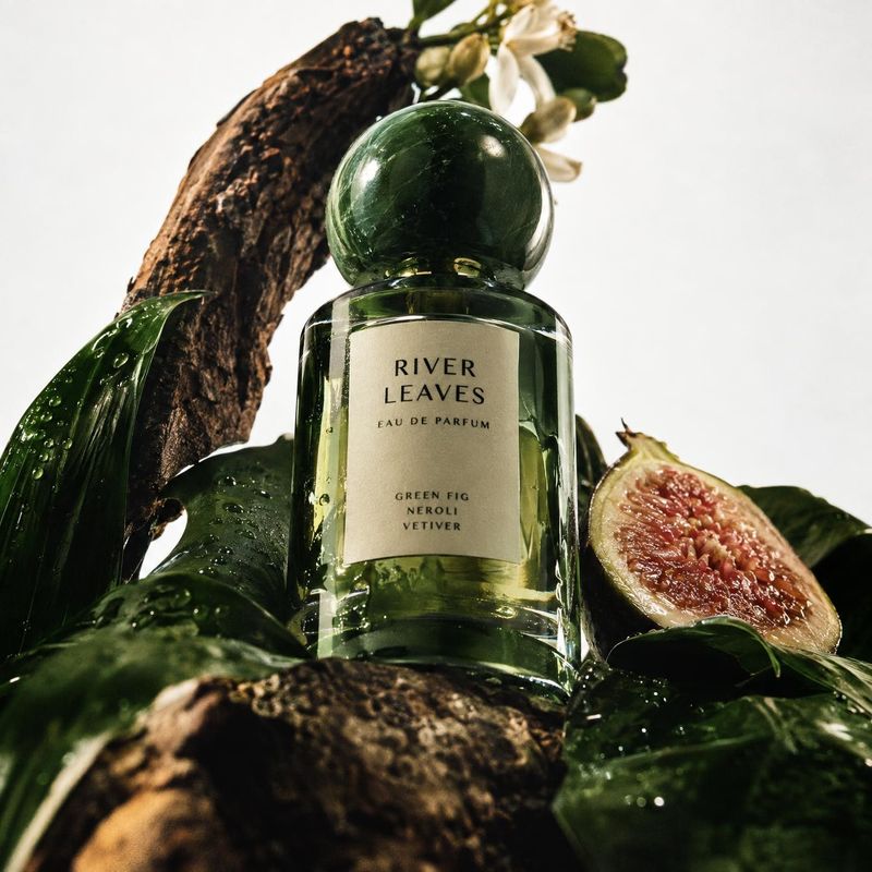 perfume ingredients product photo: A green glass perfume bottle sits among glossy leaves, bark, and a halved fig against a pale studio backdrop, creating a lus…