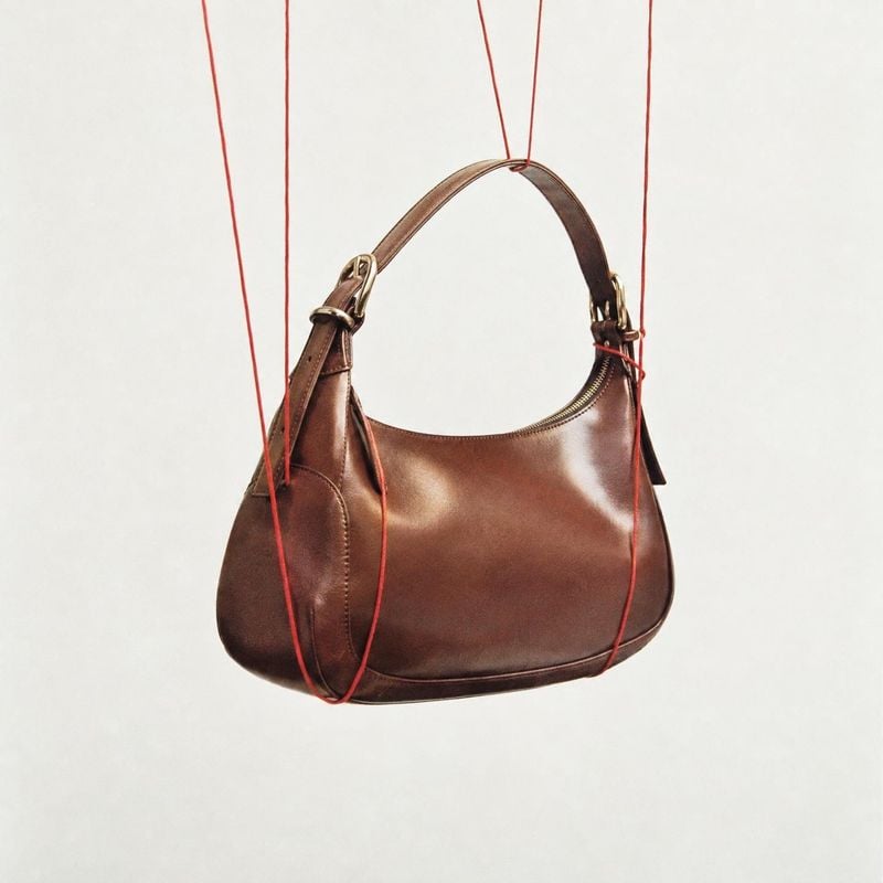 brown leather shoulder bag floating product product photo: A polished brown leather shoulder bag hangs from fine red cords against a warm off-white studio back…