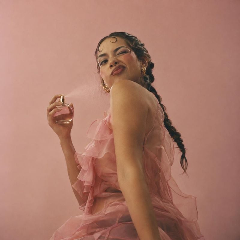 perfume product photo: A woman in a flowing pink ruffled dress poses against a matching studio backdrop while holding a perfume bottle, with soft warm light cr…
