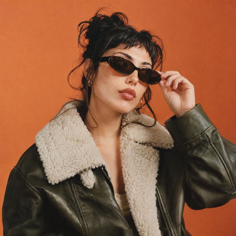 sunglasses on-body detail product photo: A woman in dark sunglasses and a shearling-collar leather jacket poses against a warm orange studio backdrop, creating…