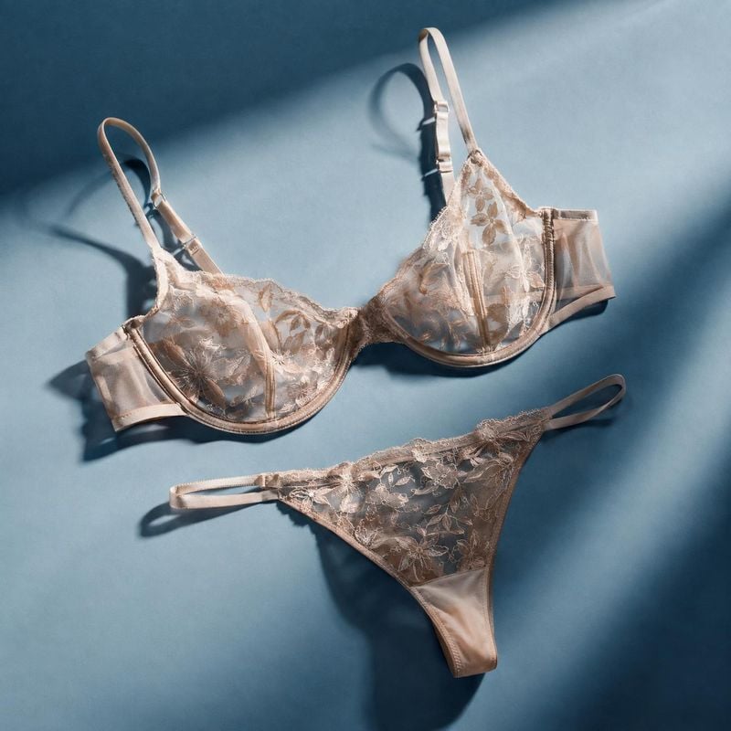 lace lingerie set flat lay product photo: A blush-toned lace bra and matching briefs are arranged on a cool blue surface, with directional light casting soft d…