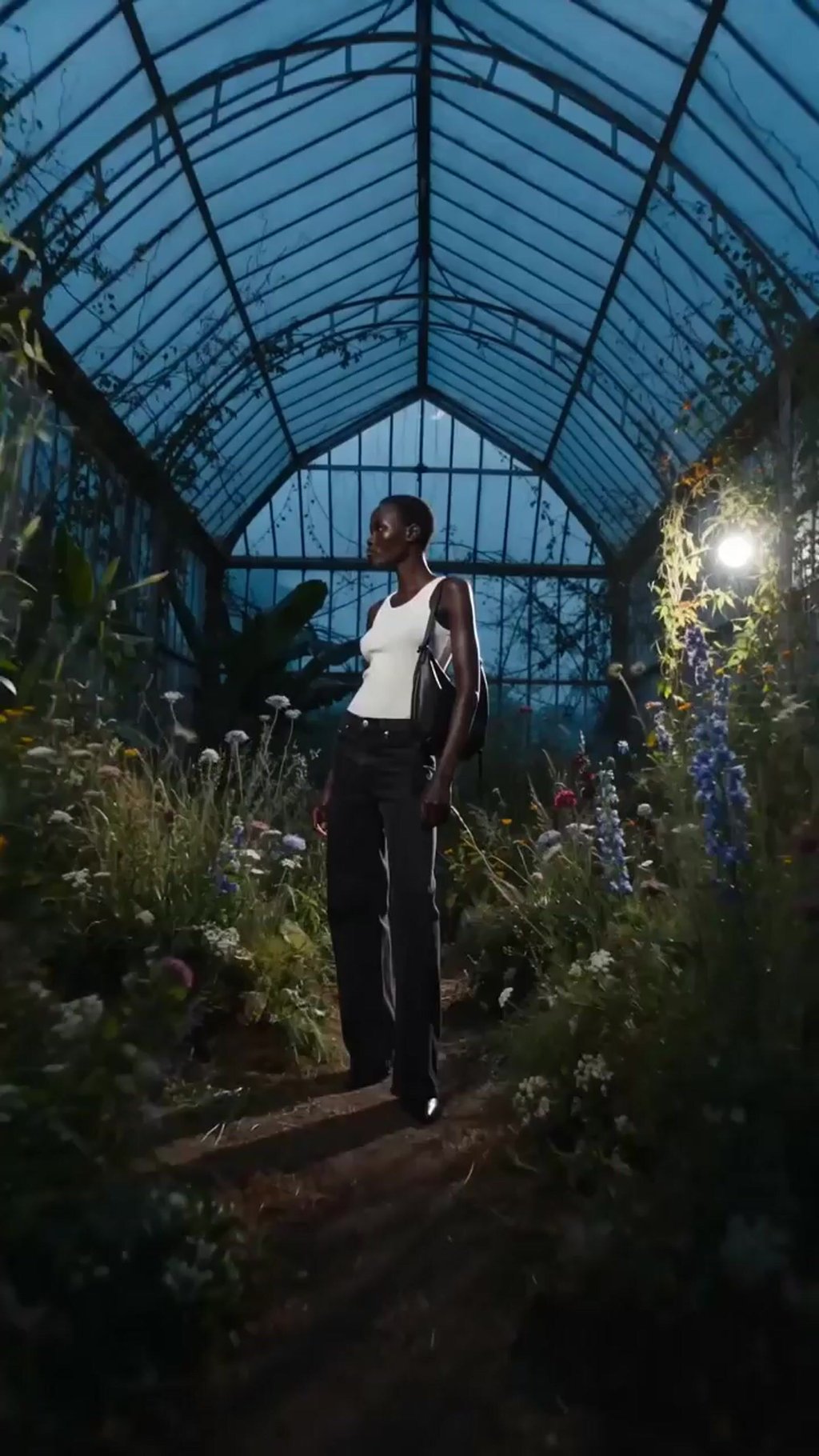 Generated model turning to camera with a black shoulder bag inside a glasshouse at dusk