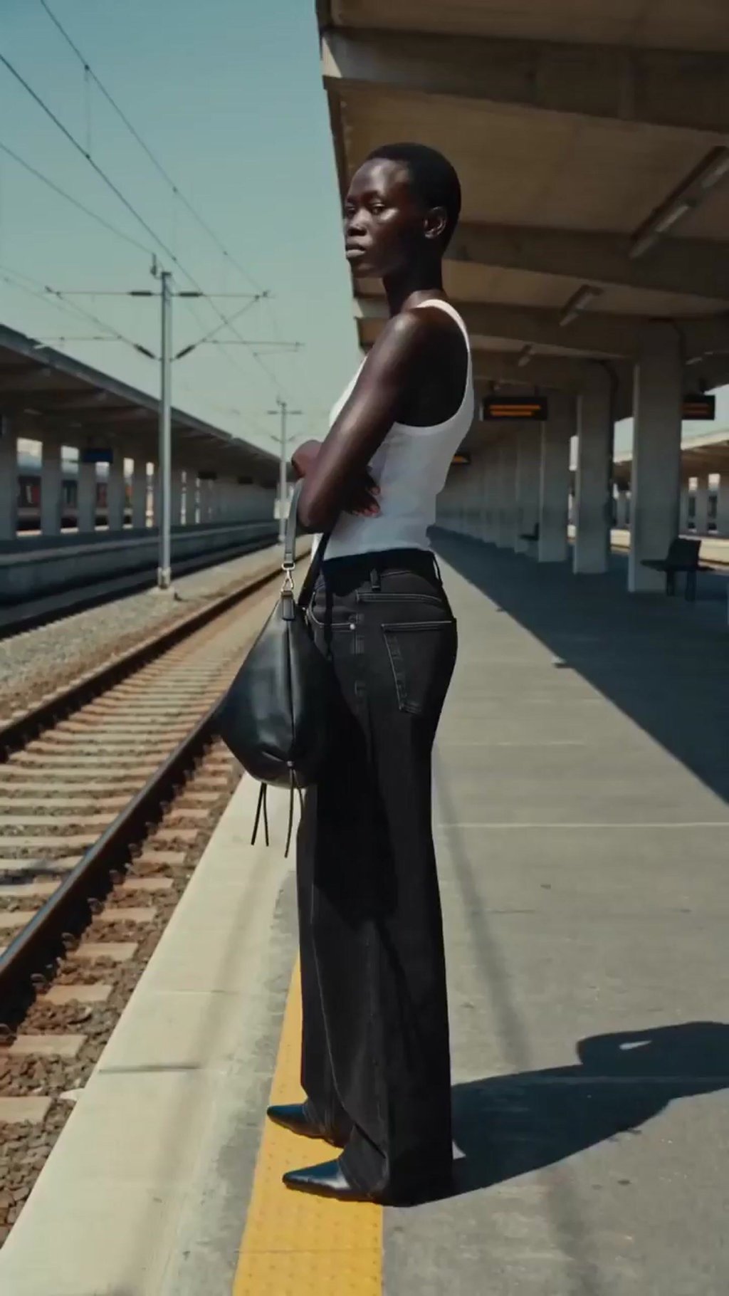 Generated model standing on a sunlit train platform with the same black shoulder bag