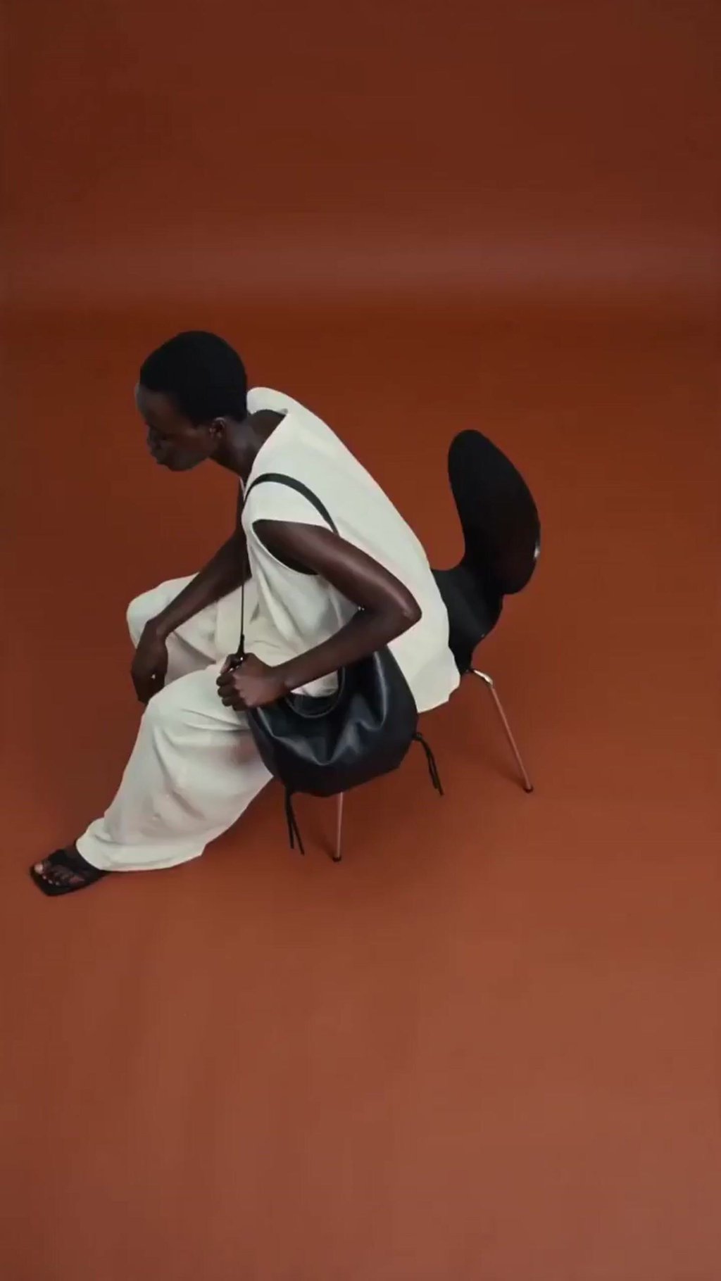 Generated model seated on a black chair against a rust-red studio wall, holding the same black shoulder bag