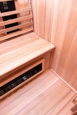 Interior of a compact sauna with pale cedar plank walls, tiered wooden benches, and black slatted heater grilles, photographed in warm light.