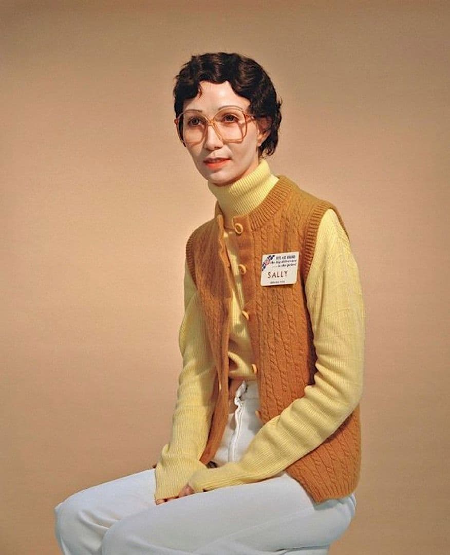 a woman sitting on a stool wearing a sweater and glasses