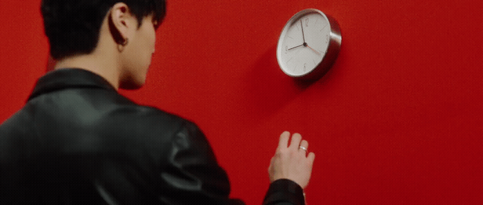 a man in a black leather jacket standing next to a red wall with a clock