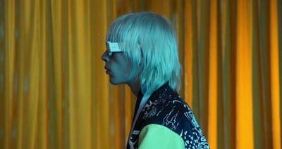 a woman with blue hair and glasses standing in front of a yellow curtain
