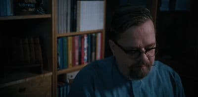 a man with glasses and a beard sitting in front of a book shelf