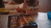 a little girl sitting at a table painting with a brush