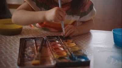 a little girl sitting at a table painting with a brush