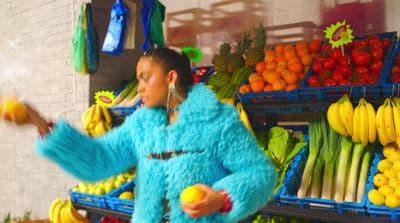 a woman in a blue fur coat standing in front of a fruit stand