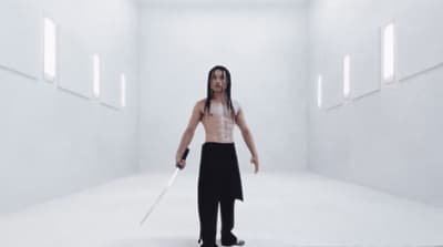 a man with long hair holding a sword in a white room