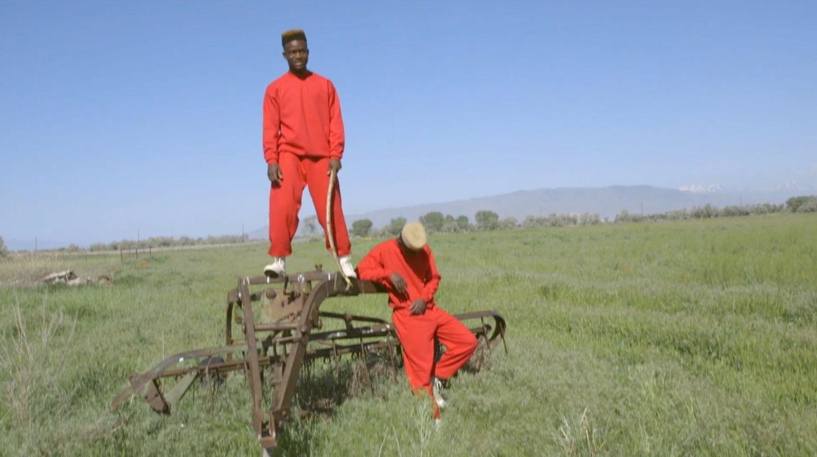 two men in red jumpsuits standing on a cart in a field