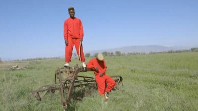 two men in red jumpsuits standing on a cart in a field