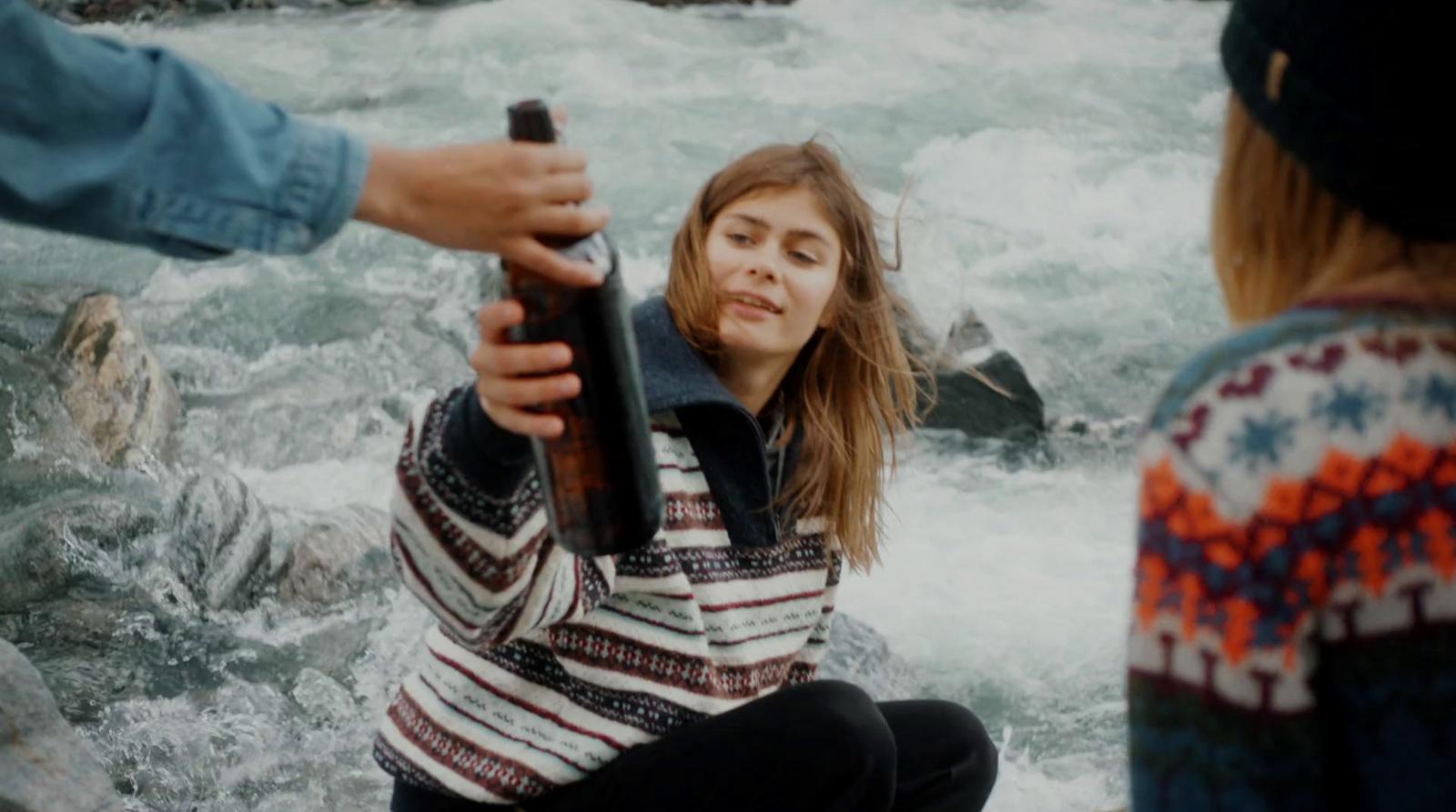 a woman is holding a beer bottle in front of a river
