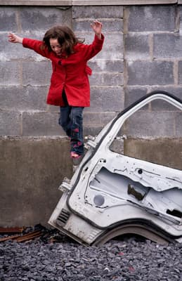a girl in a red jacket jumping over a broken car
