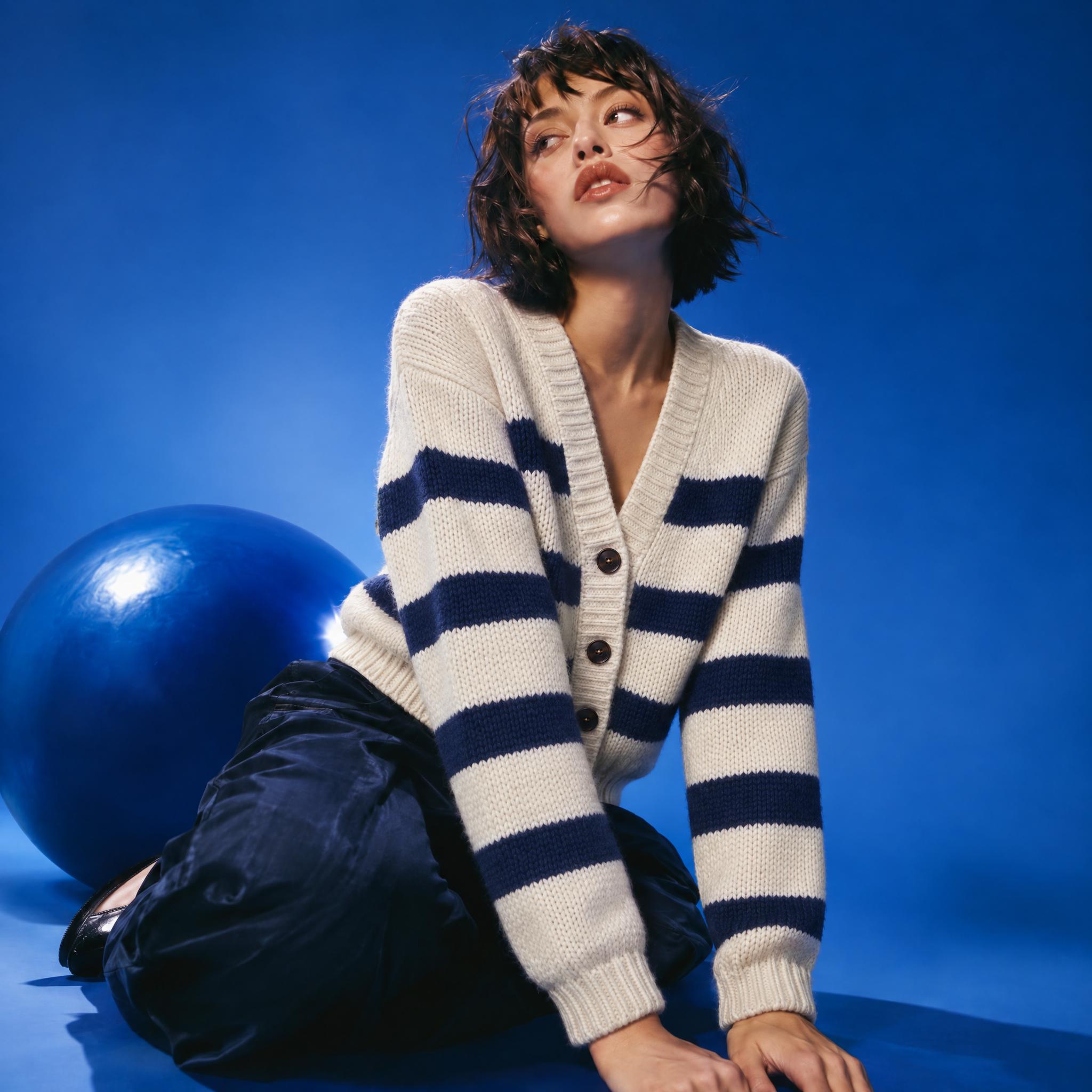 Cream cardigan with navy stripes laid flat on gray, and the same cardigan worn by a seated model in front of a blue backdrop and a blue ball - After