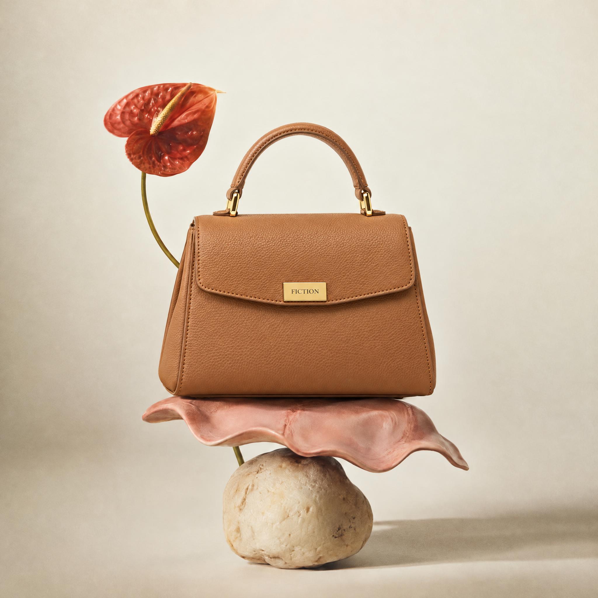 Tan leather top-handle bag on a cream backdrop, and the same bag balanced on a pink shell and a pale stone beside a red anthurium - After