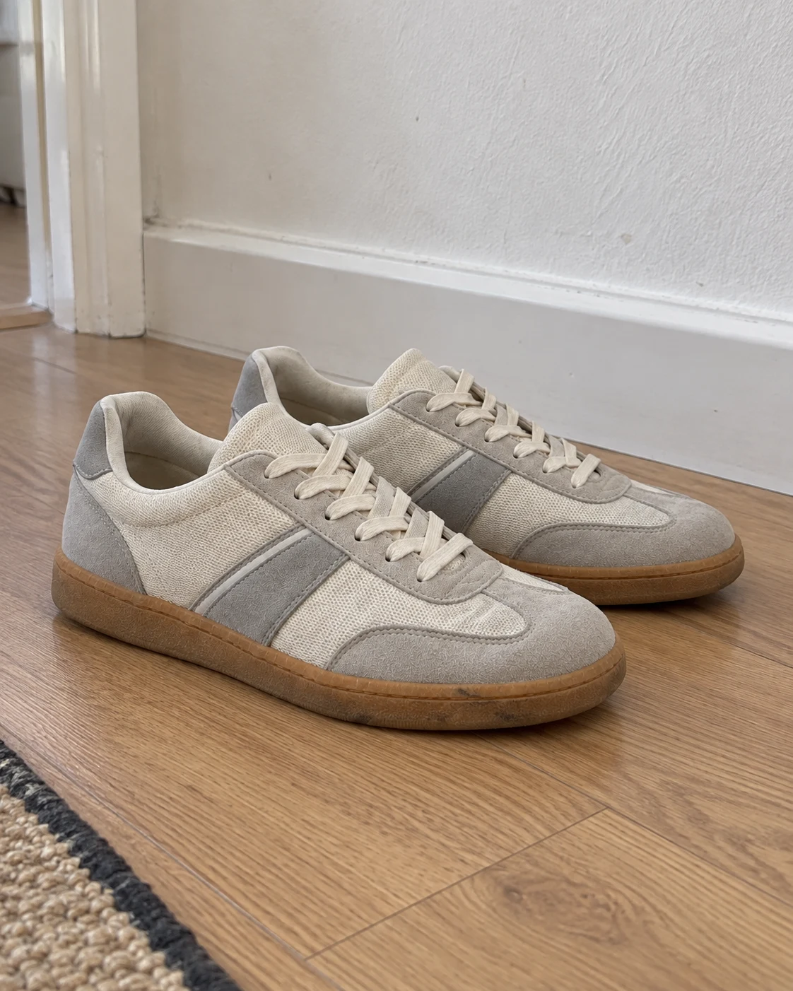 Lightly worn sneakers on a wooden hallway floor beside a white wall and rug